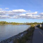 Paved riverside pathway beside the wide Bow River under puffy clouds, with the downtown Calgary skyline in the distance