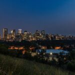 Downtown Calgary skyline lit up at blue hour under a deep blue sky, seen from a grassy hillside viewpoint with a river and bridge at right