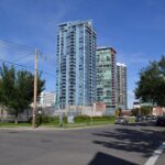 Glass high-rise condominium towers on a sunny tree-lined Beltline street corner, with the Calgary Tower in the background